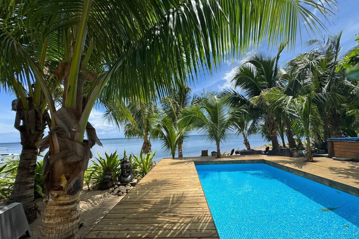 A wooden deck leading to a pool with a clear blue water surface, surrounded by palm trees and a view of the ocean.