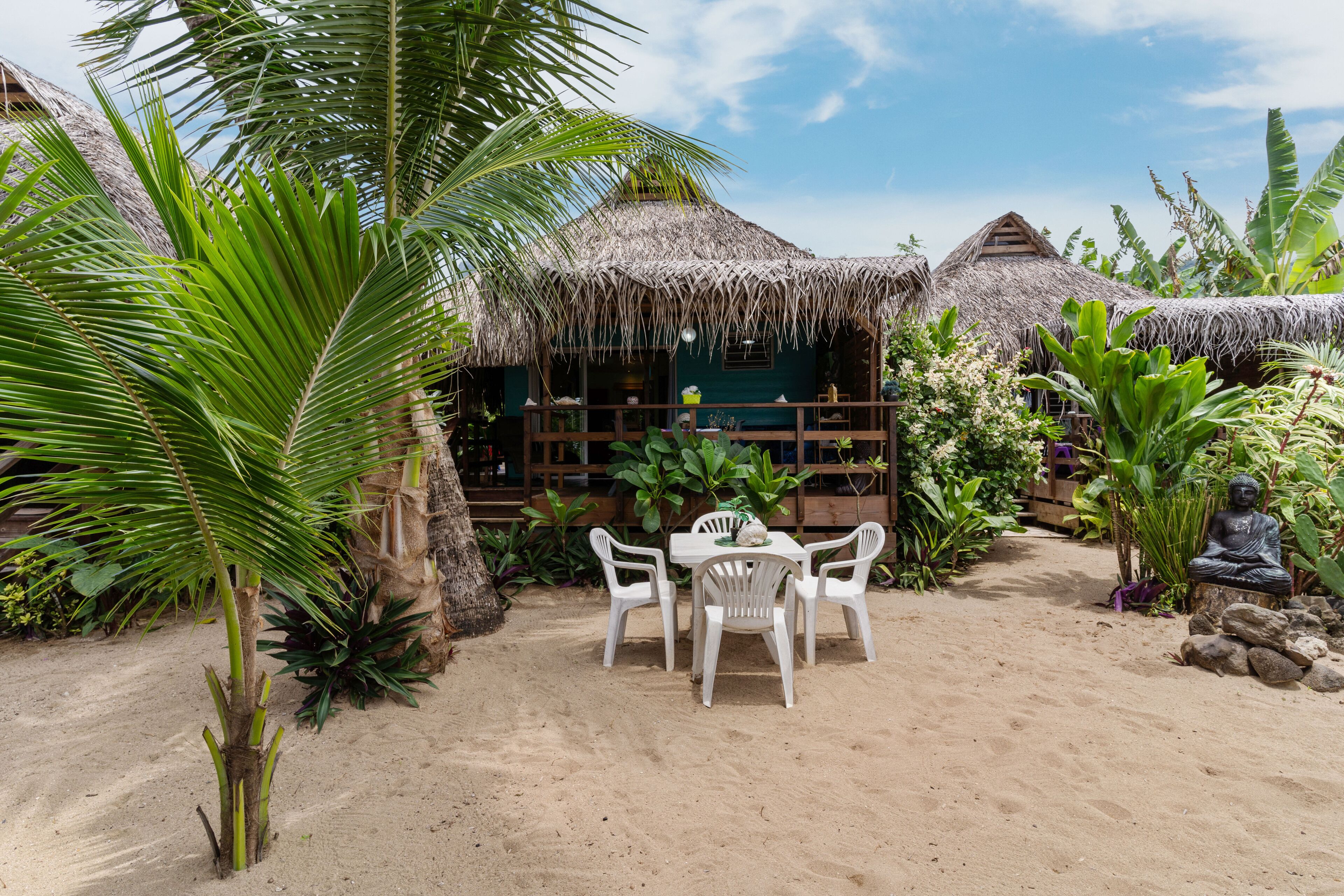 A beachside resort with thatched-roof huts, a sandy patio with a table and chairs, and tropical vegetation.