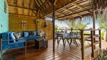 Eine Holzterrasse mit einem Reetdach, einem blauen gemusterten Sofa und einem Tisch mit Stühlen.