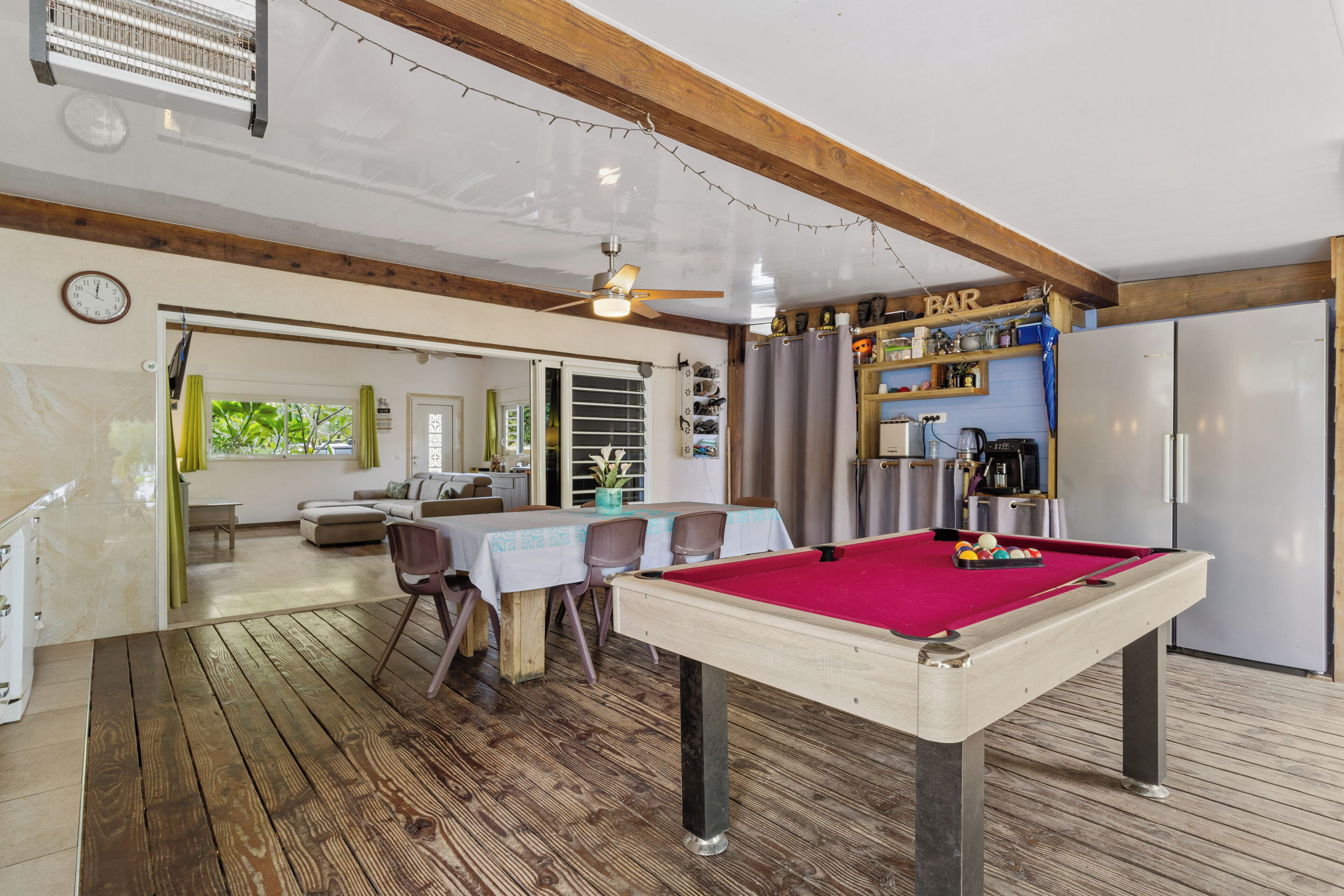 A pool table in a room with wooden floors, a ceiling fan, and a bar area with shelves.
