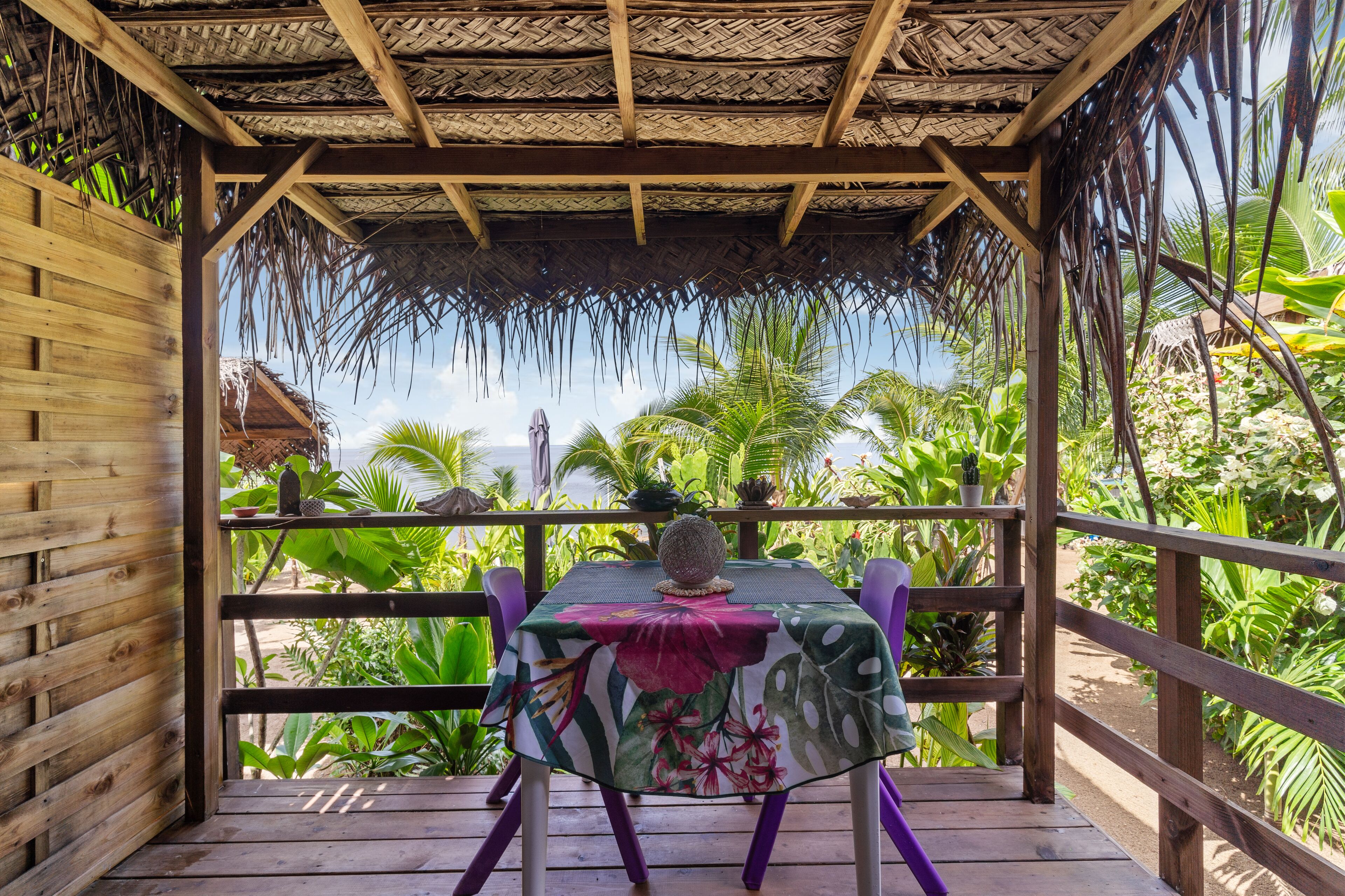 Una terraza de madera con una mesa y sillas, rodeada de exuberante vegetación tropical y una estructura de techo de paja.