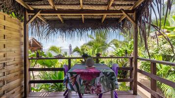 Una terraza de madera con una mesa y sillas, rodeada de exuberante vegetación tropical y una estructura de techo de paja.