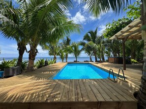 Um deck de madeira com piscina, rodeado por palmeiras e vista para o oceano.