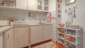 A kitchen with light wooden cabinets, a tiled backsplash, and a red-tiled floor. There is a sink, a countertop with kitchen utensils, and a shelf with various items. - Casa Celeste in Pomonte (Marciana)