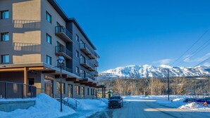 A modern multi-story building with balconies, a snow-covered road, and a mountain range in the background. - Modern 2BR in Golden w/ Stunning  MTN Views! (Golden)