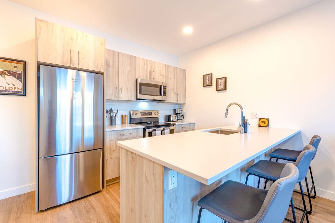 Una cocina moderna con refrigerador de acero inoxidable, microondas y estufa, gabinetes de madera, una isla de encimera blanca con taburetes de bar y un fregadero.