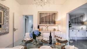Un espace repas moderne avec une table en verre, des chaises blanches, un lustre et un élément décoratif mural.