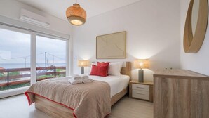 Une chambre moderne avec un grand lit, deux tables de chevet, une petite table en bois, une grande fenêtre avec vue et un luminaire au plafond.