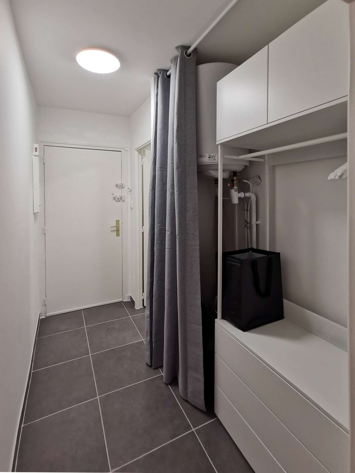 Un couloir avec une porte blanche, des rideaux gris, un sol carrelé et un meuble de rangement blanc sur lequel repose un sac noir.