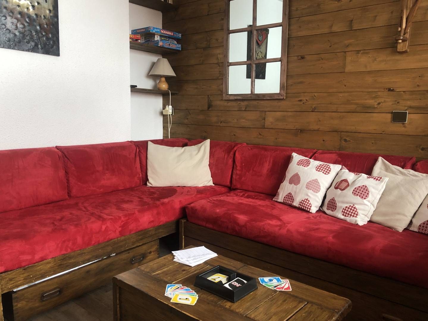 Un canapé d’angle rouge agrémenté de coussins à motifs, une table basse en bois avec un jeu de cartes, et un mur en bois orné d’un miroir et d’étagères.
