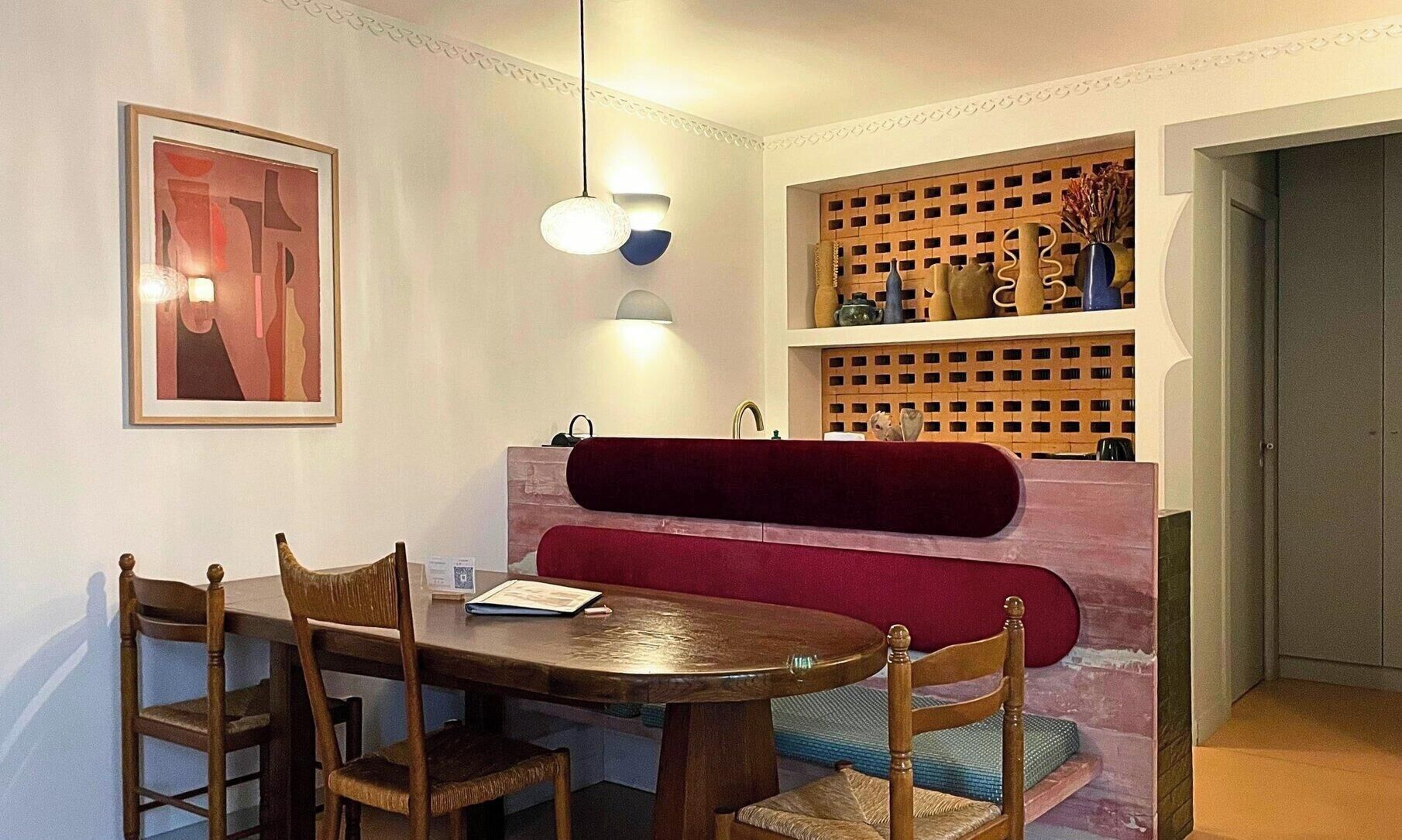 Een eethoek met een houten tafel en stoelen, een rode, beklede bank en een wandplank met decoratieve objecten.