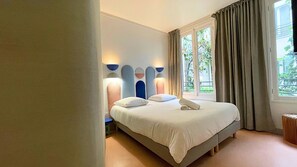 Ein Schlafzimmer mit einem Bett, Nachttischlampen, einem Fenster mit Vorhängen und Blick ins Grüne.