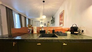 Et moderne køkken med en sort vask, kaffemaskine og elkedel. Der er et vindue med gardiner, en vase med blomster og en vægmonteret lampe.