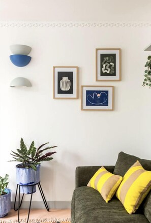 Un salon avec un canapé gris, des coussins jaunes et gris, une plante bleue en pot et trois œuvres d’art encadrées accrochées au mur.