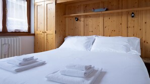 Un letto rifatto con lenzuola bianche e asciugamani piegati, pannelli di legno sulle pareti e un piccolo ripiano ricoperto da un tessuto a quadri.