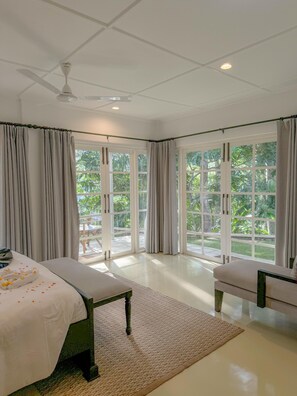 Habitación de hotel con dos camas, ventilador de techo, ventanas amplias y vista a follaje exterior.