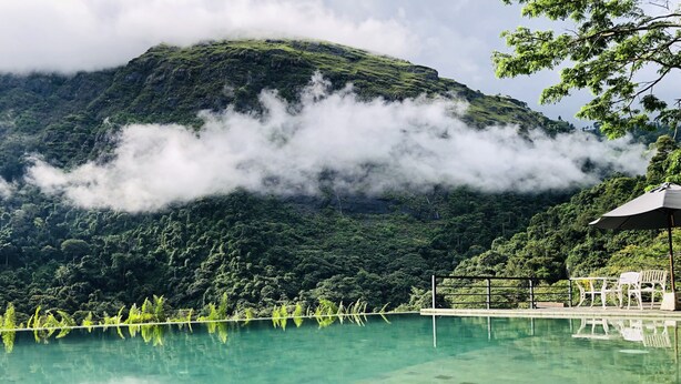 Une piscine aux eaux cristallines, avec une chaîne de montagnes en toile de fond, entourée d’une végétation luxuriante et agrémentée d’un belvédère avec une table et des chaises.