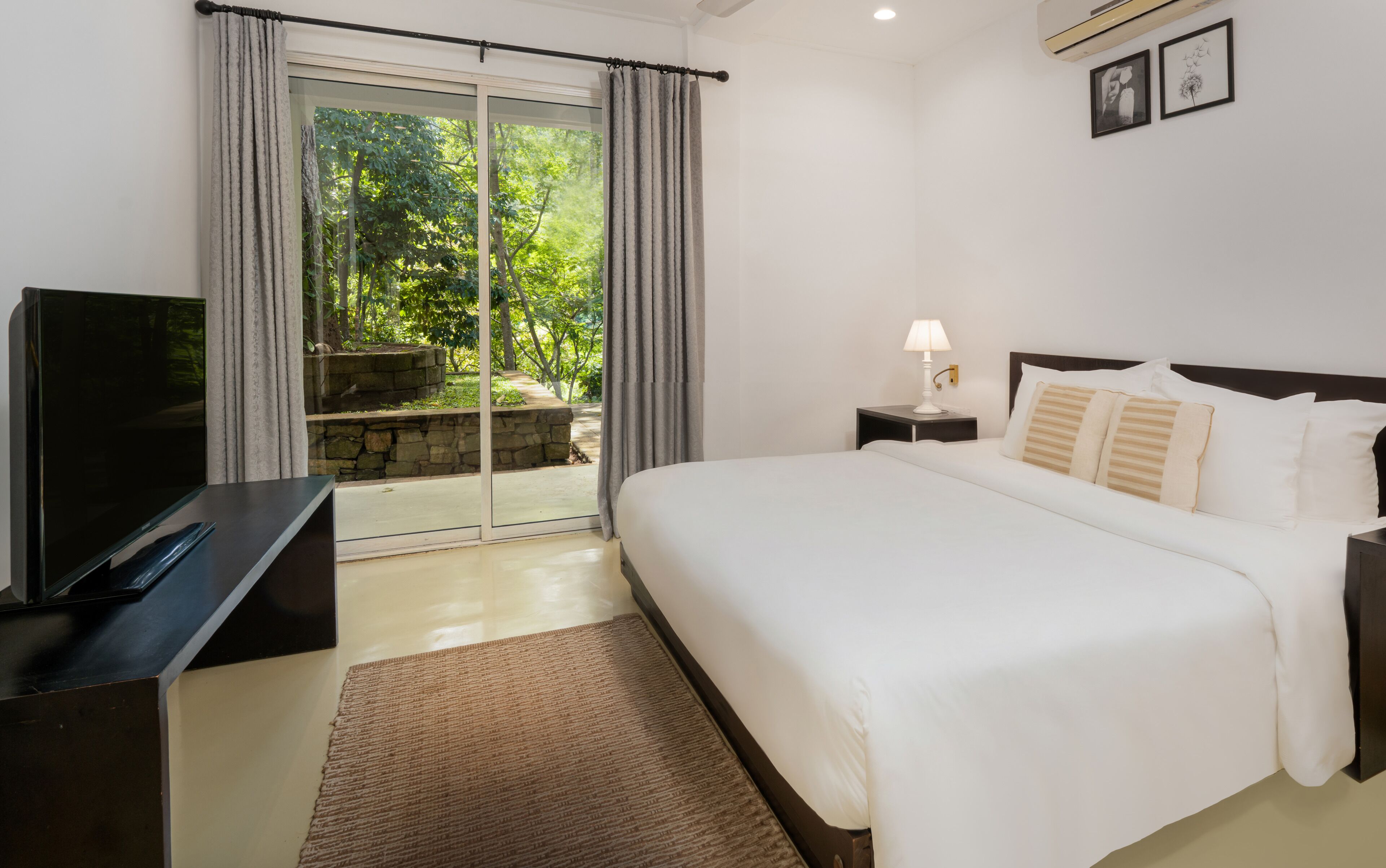 Quarto de hotel com uma cama, televisão, mesa de cabeceira, luminária e vista para um jardim através de uma porta de vidro.