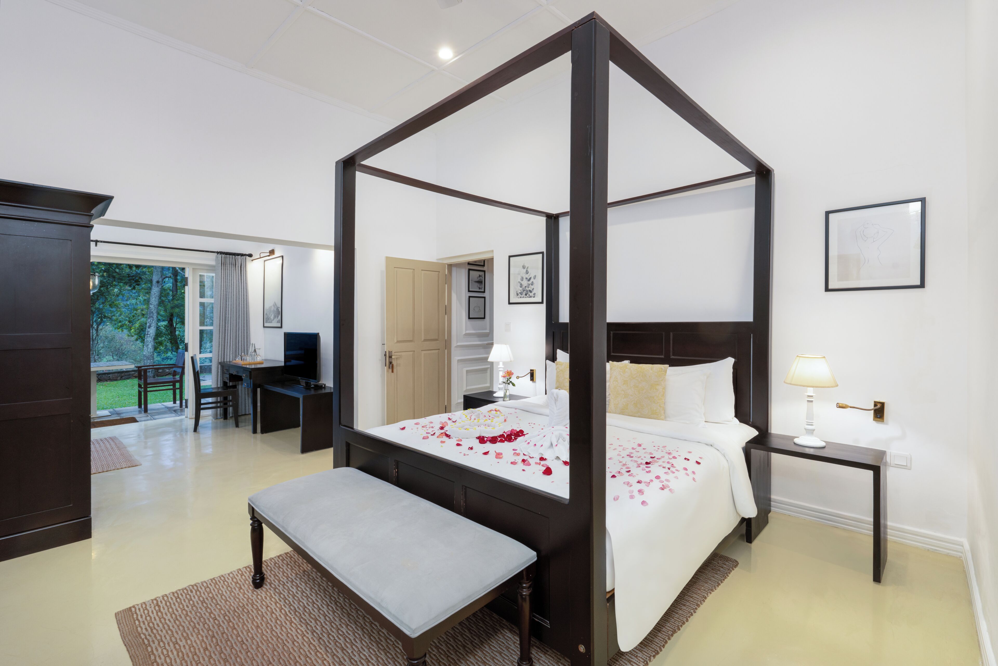 A four-poster bed with rose petals, a desk, and a bench in a hotel room.
