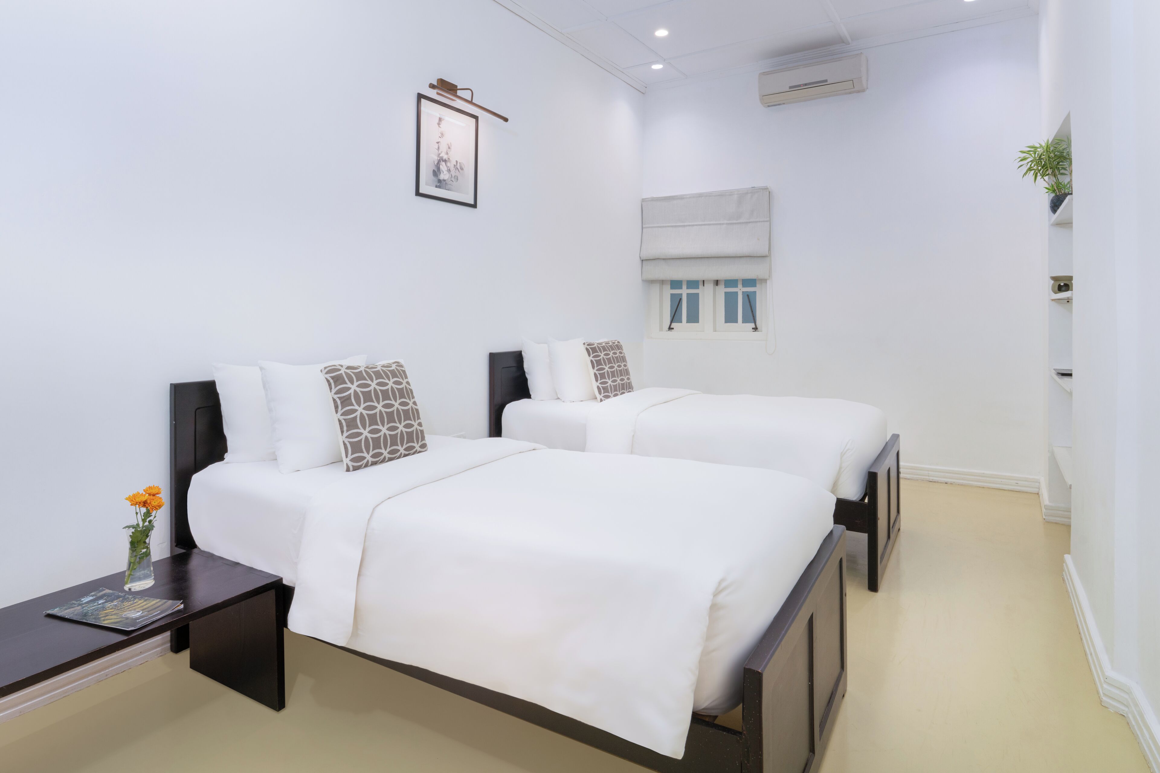 A hotel room with two beds, a nightstand, a framed picture on the wall, an air conditioning unit, and a window with a blind.