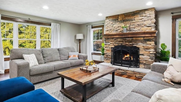 A living room with a stone fireplace, a grey sofa, a coffee table, and large windows with curtains.