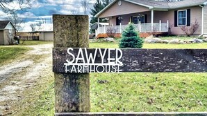 A wooden sign with 'SAWYER FARMHOUSE' written on it, a house with a porch, and a clear sky.