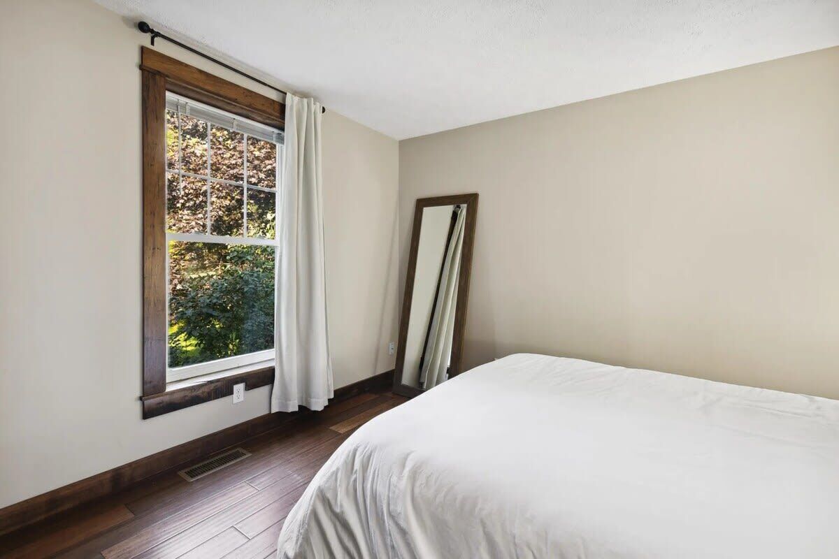 A bedroom with a bed, a window with curtains, a mirror, and a view of trees.