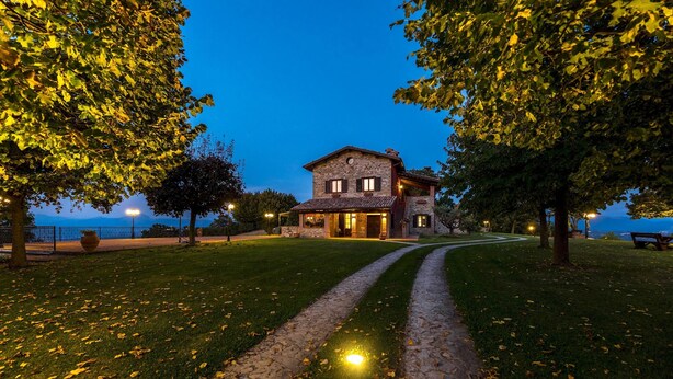A stone house with a paved path leading to it, surrounded by trees and a body of water in the background. - Tenuta Montiepoli 12 3 Emma Villas (Valfabbrica)
