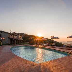 A pool area with lounge chairs and umbrellas, a stone building, and a sunset in the background. - Tenuta Montiepoli 12 3 Emma Villas (Valfabbrica)