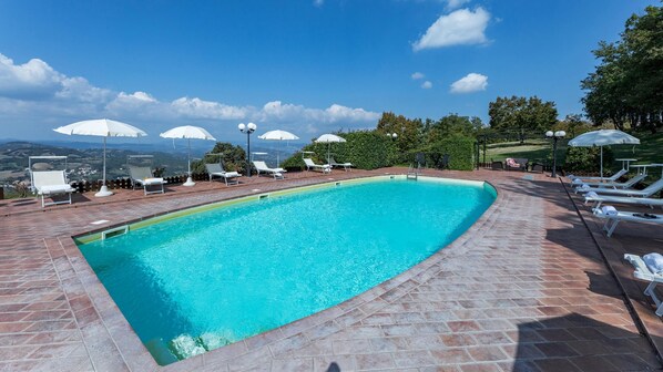 A swimming pool with sun loungers and umbrellas, surrounded by a brick patio and a view of the countryside. - Tenuta Montiepoli 12 3 Emma Villas (Valfabbrica)