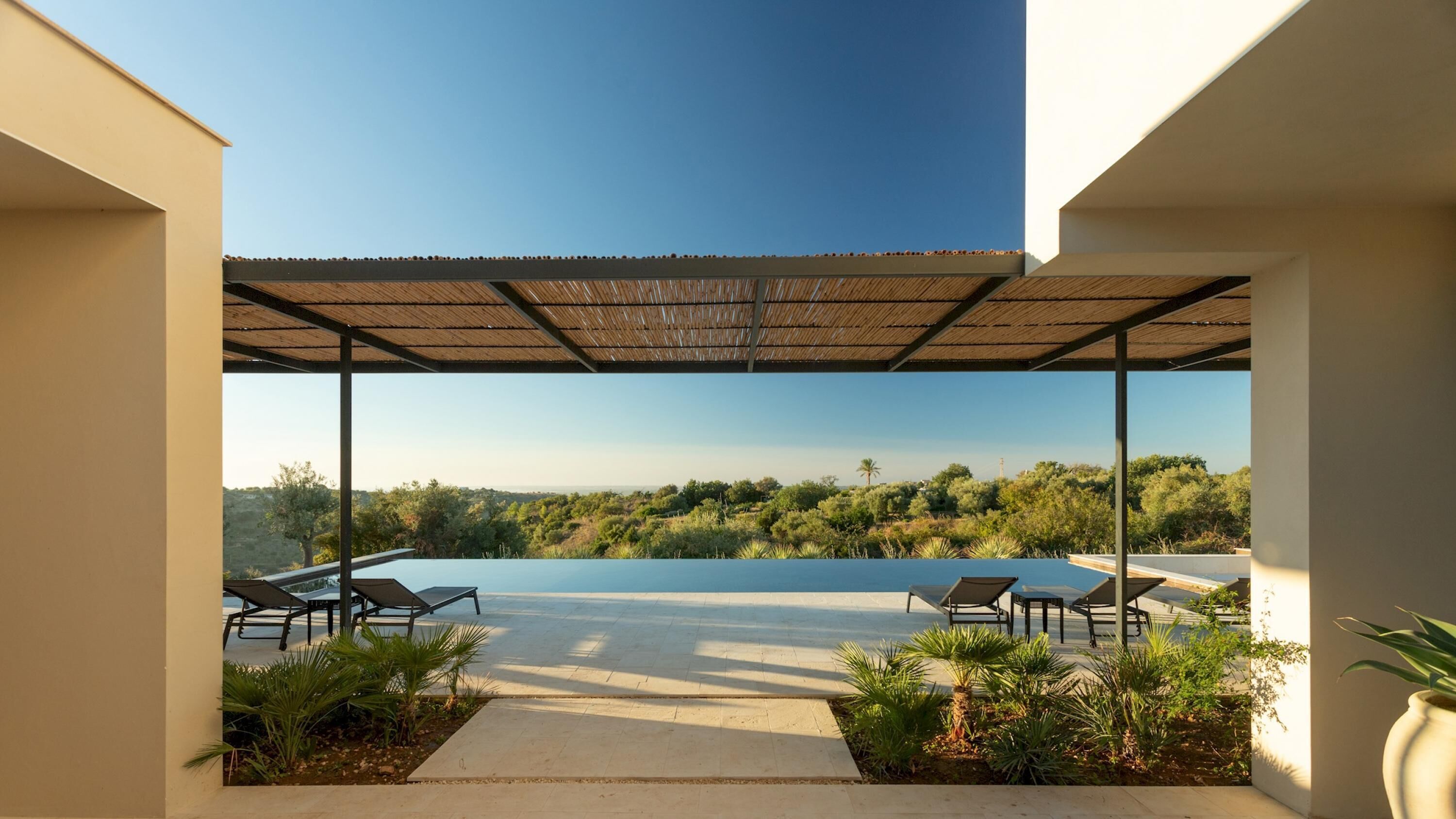 An infinity pool with lounge chairs under a pergola, surrounded by greenery and a view of a forested landscape.