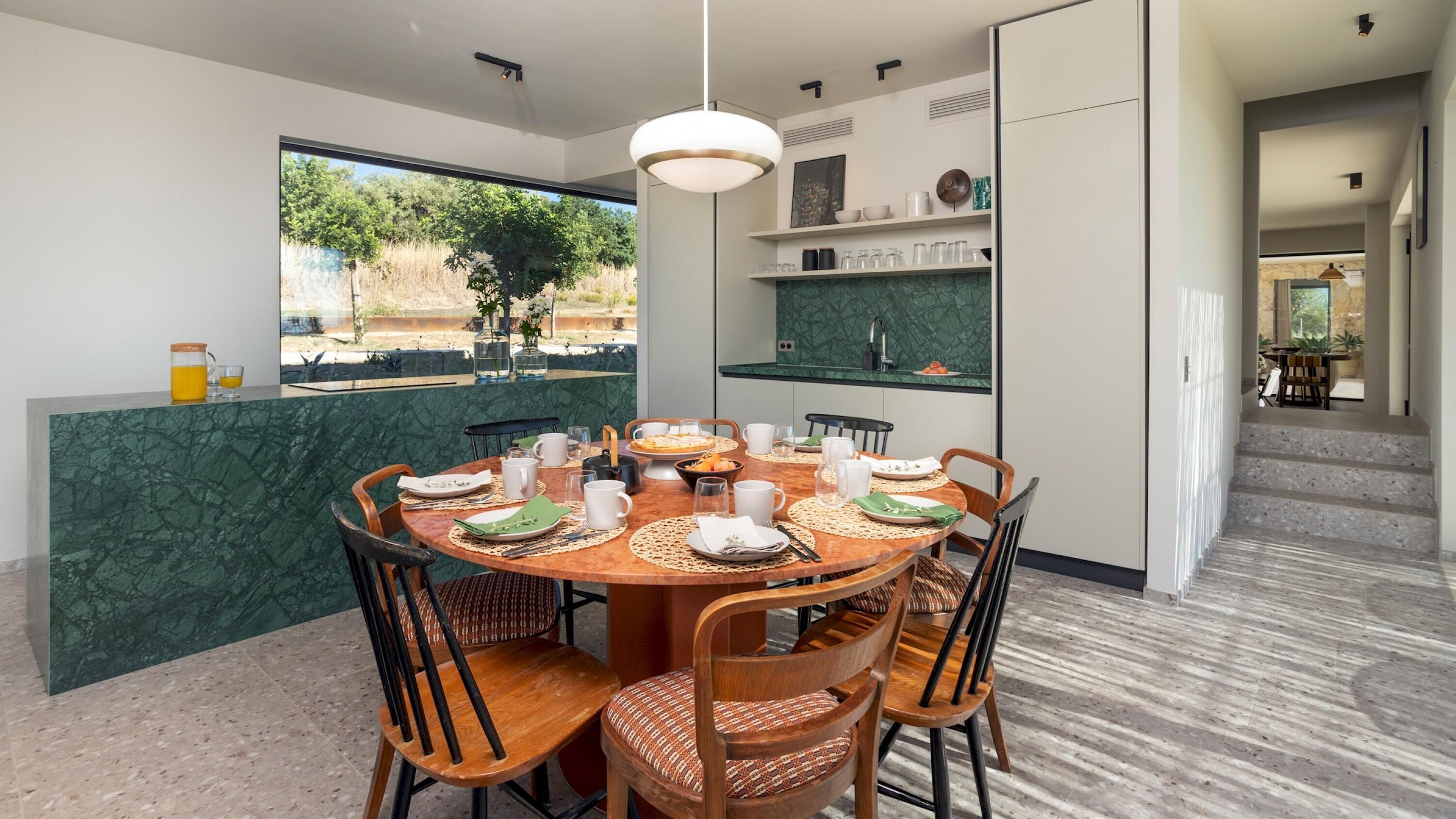 A modern dining area with a round table set for four, surrounded by wooden chairs with woven seats. A green marble countertop with a sink is visible in the background.