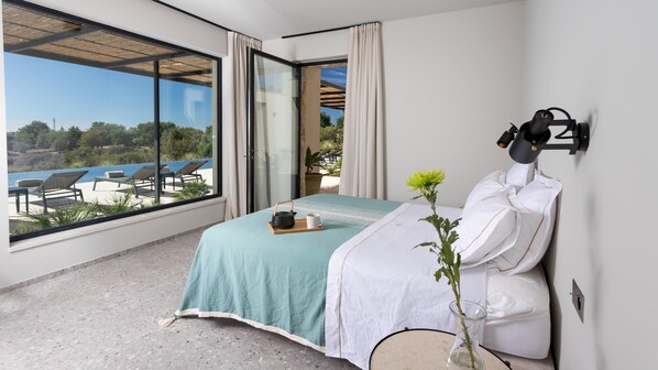 A bedroom with a large window overlooking a pool and garden, a bed with a teal bedspread, a bedside table with a kettle and mug, and a wall-mounted lamp.