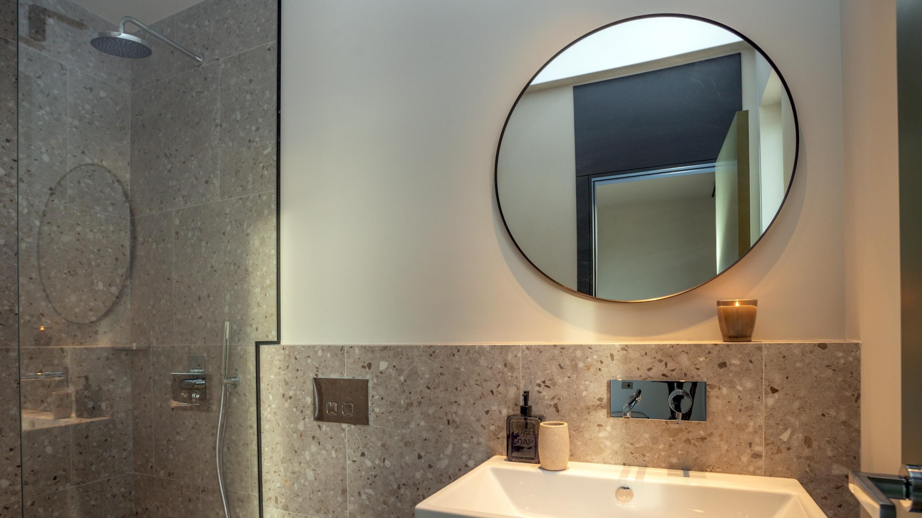 A modern bathroom with a round mirror, a sink, a handheld showerhead, and a soap dispenser.