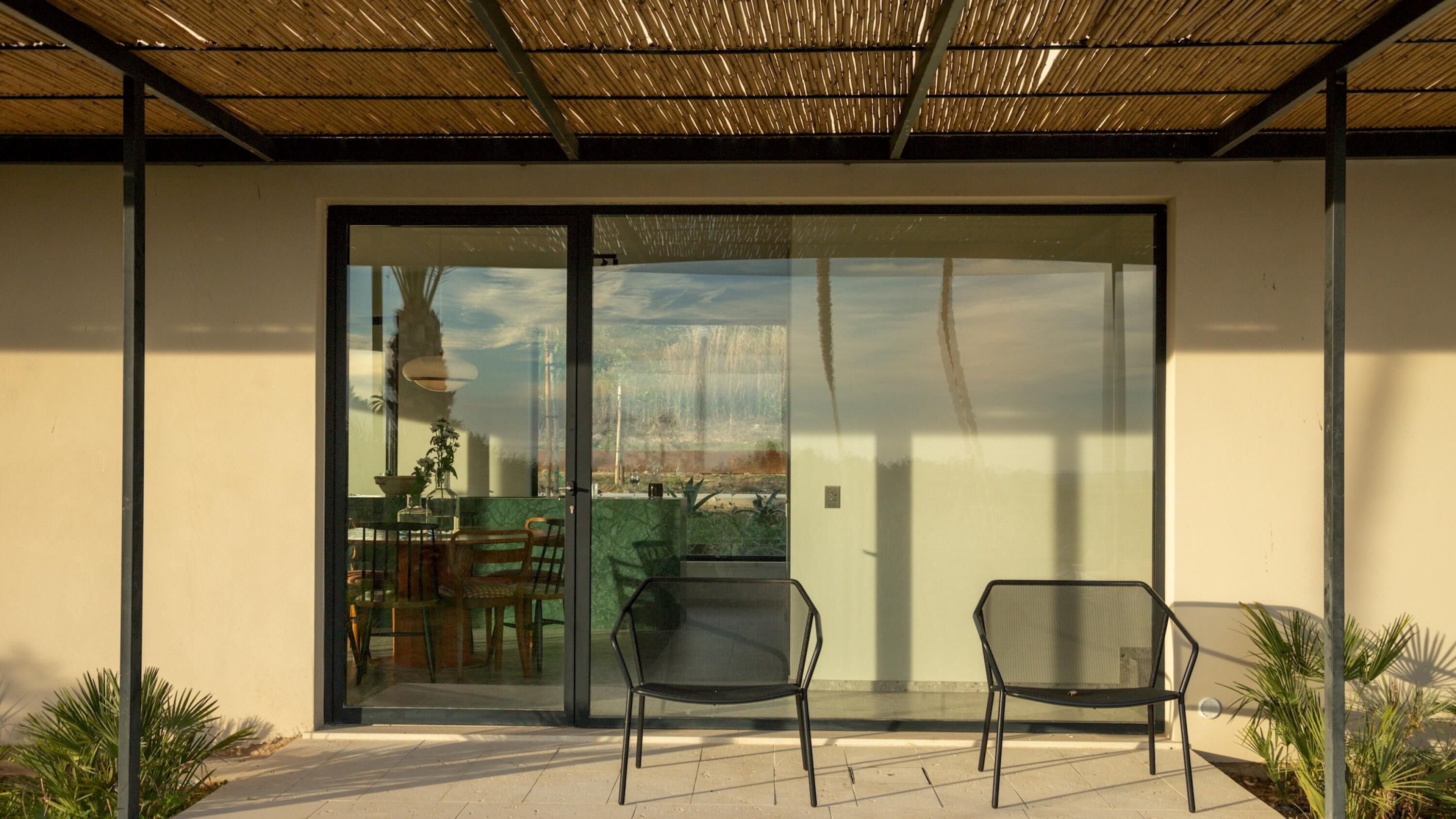 A modern patio with two chairs, a table, and a glass door leading to an outdoor area with a view of the sky and landscape.
