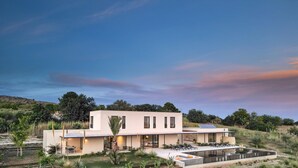 A modern two-story house with a large outdoor pool, surrounded by greenery and hills. - Villa Amore 8 Emma Villas (Avola)