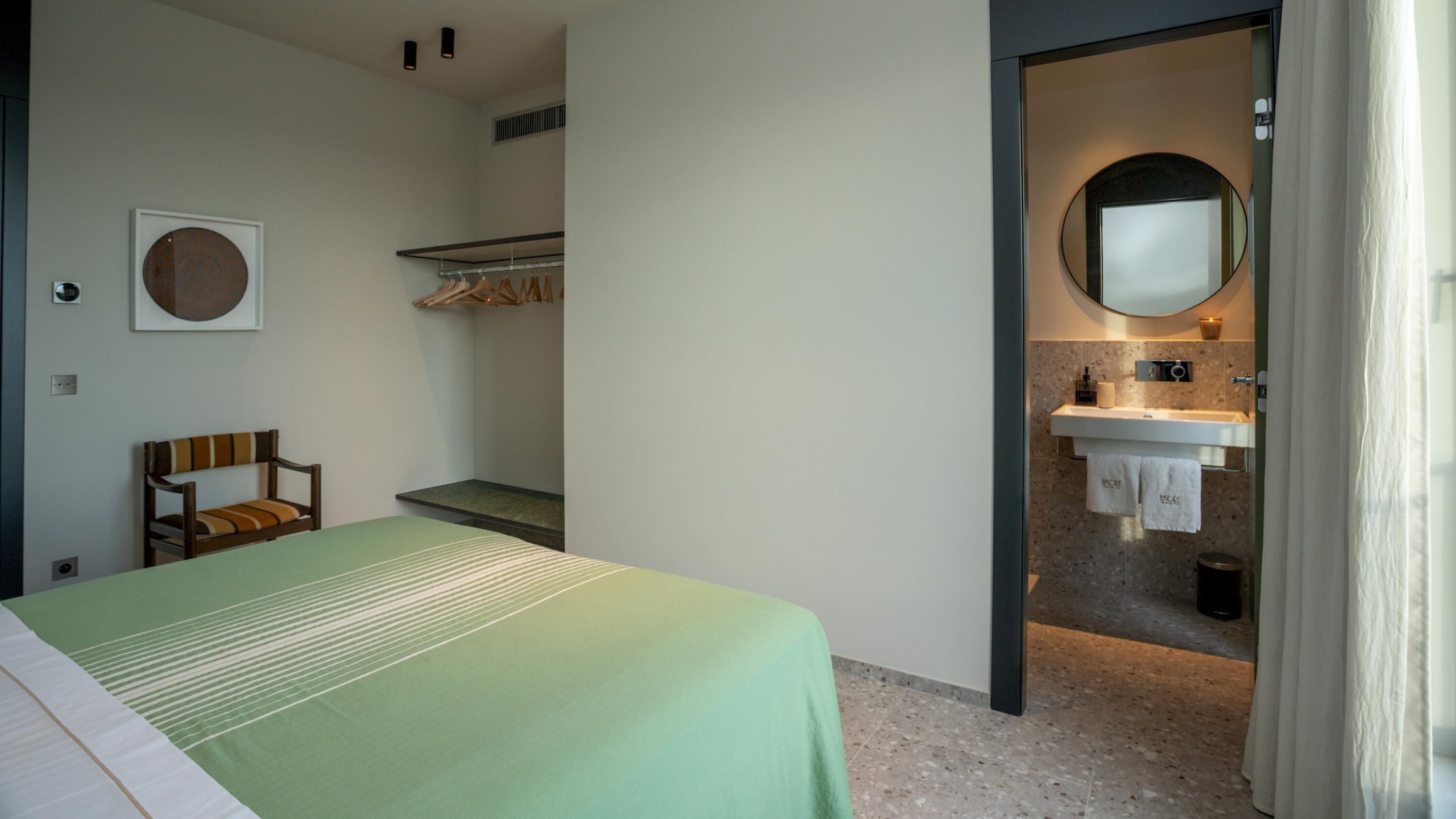Una habitación de hotel con cama, una silla, armario, espejo y baño con lavamanos e inodoro.