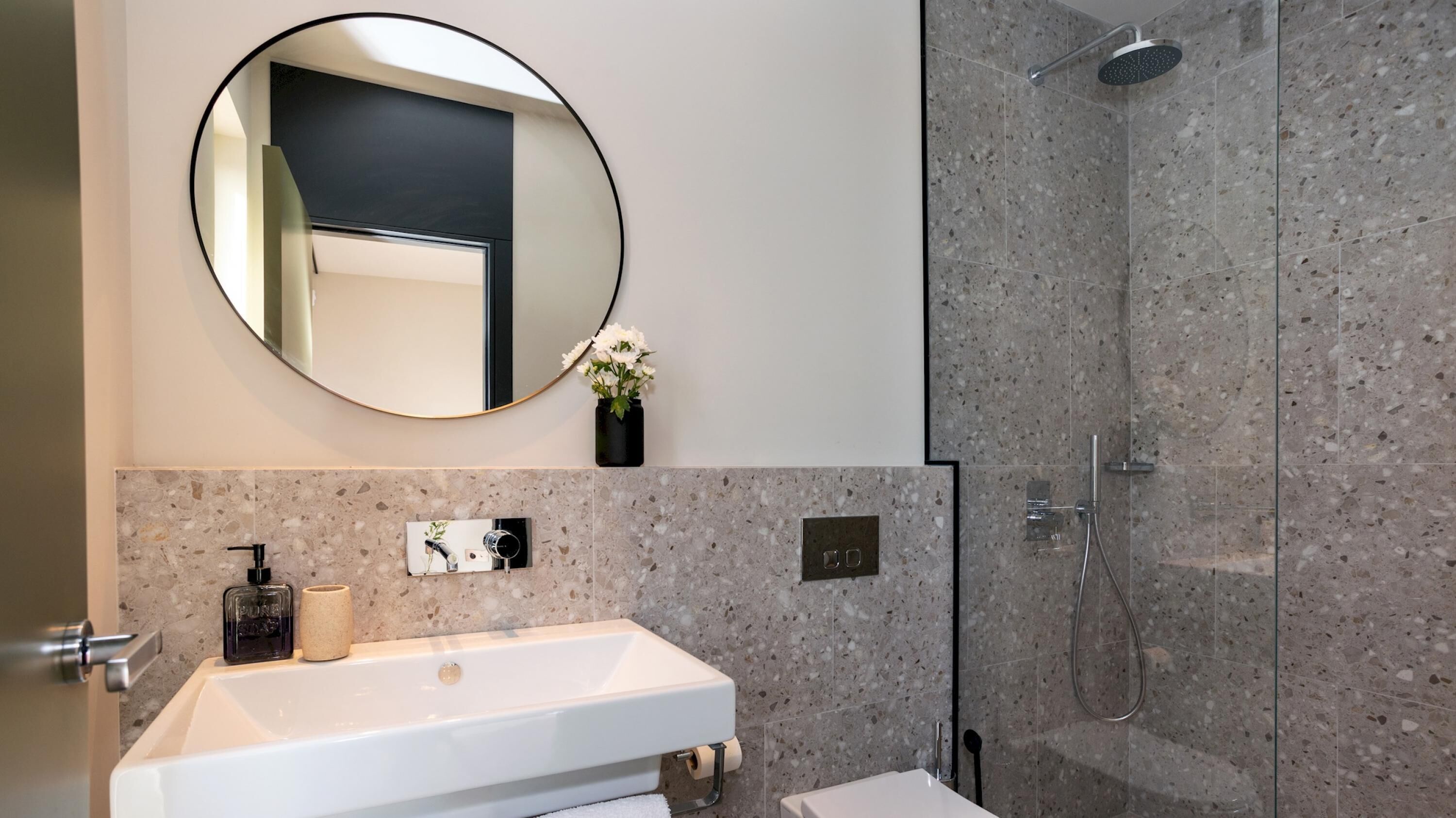 Un baño moderno con un lavamanos blanco, un espejo redondo y una zona de ducha.