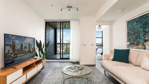 A modern living room with a television, a glass coffee table, a sofa, and a view of the cityscape through a large window. - Darlinghurst — Newly renovated 1BR + Parking (City of Sydney)