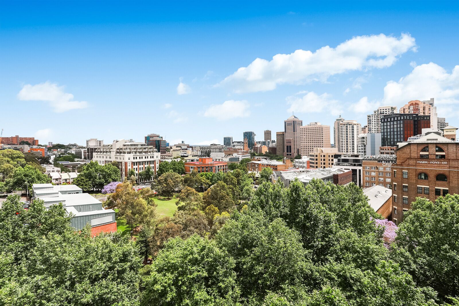 A cityscape with a mix of modern and older buildings, a prominent clock tower, and a dense tree line in the foreground.