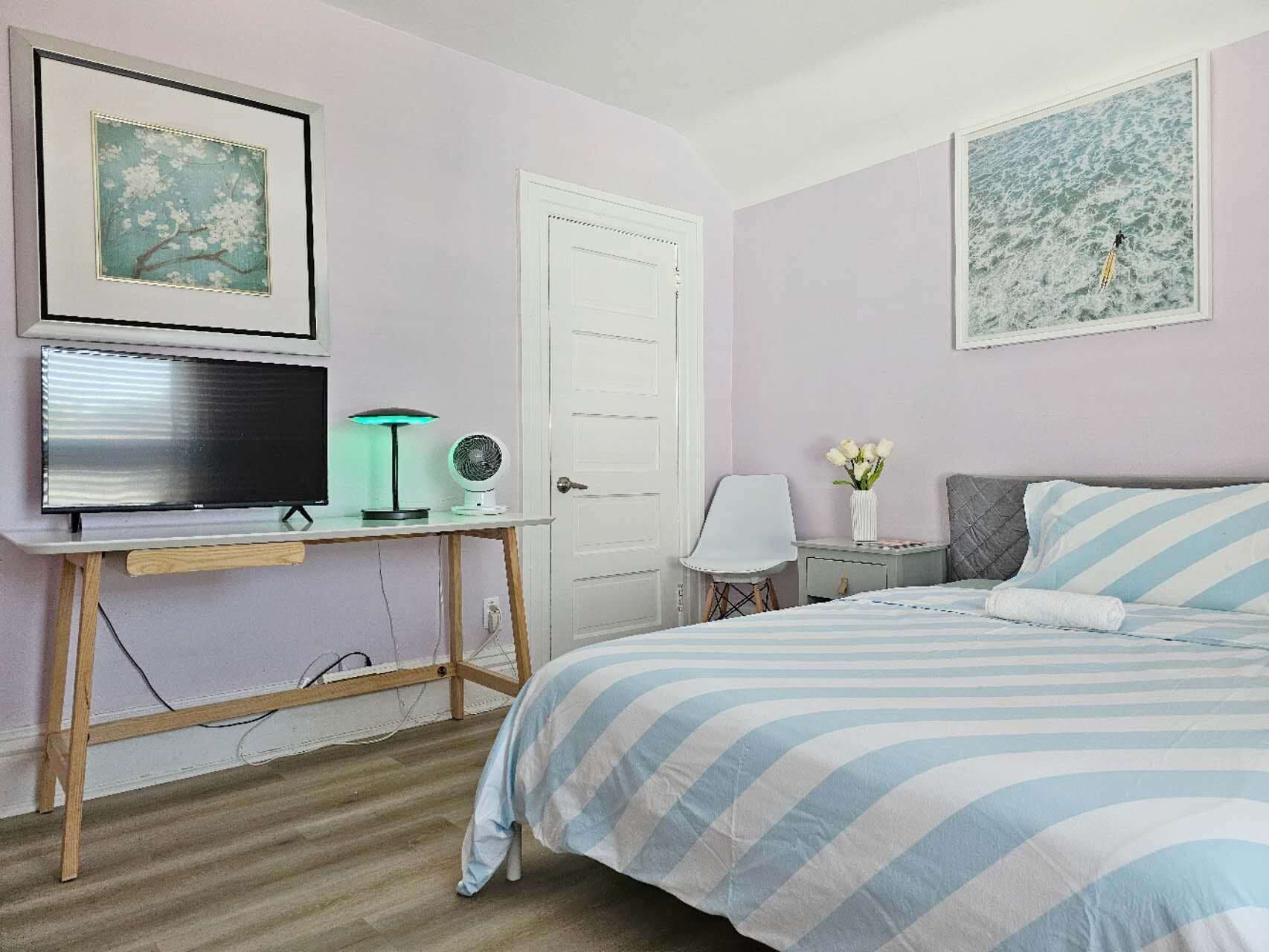 A bedroom with a bed, a desk with a computer, a chair, a small table with a lamp, and a door.