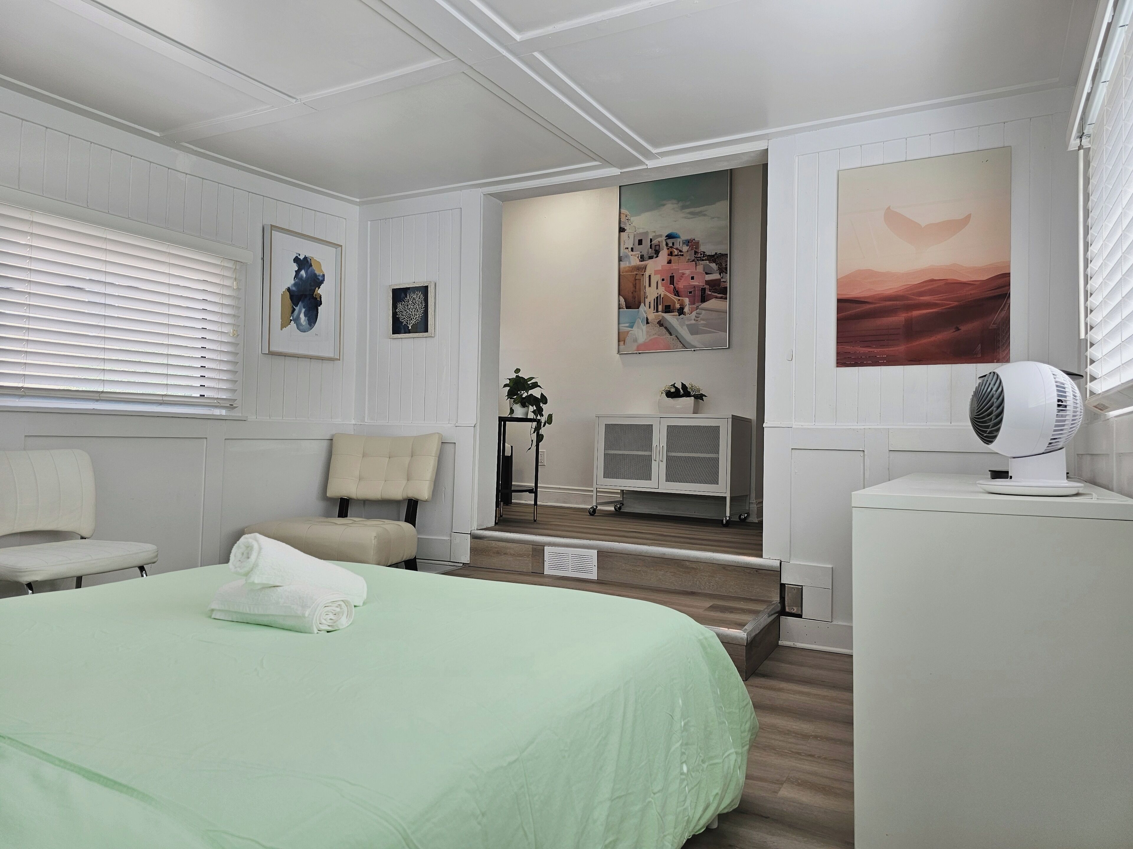 A hotel room with a bed, a chair, a desk, and a fan. There are paintings on the walls and a window with blinds.
