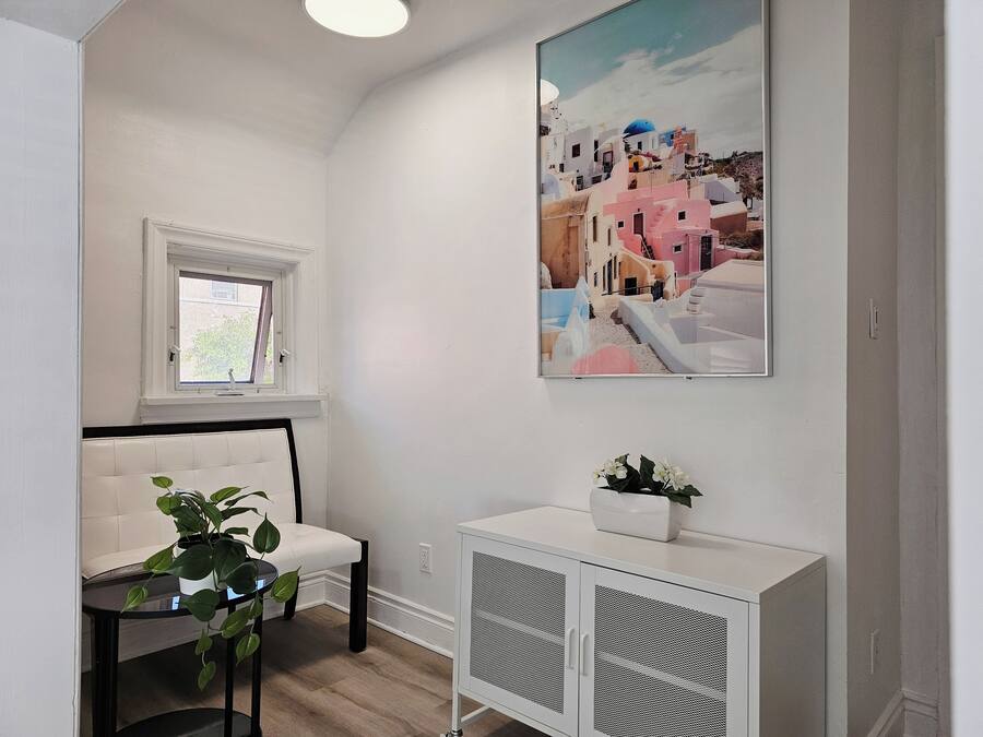 A room with a white sofa, a small table with a plant, a cabinet, and a painting of a coastal town.