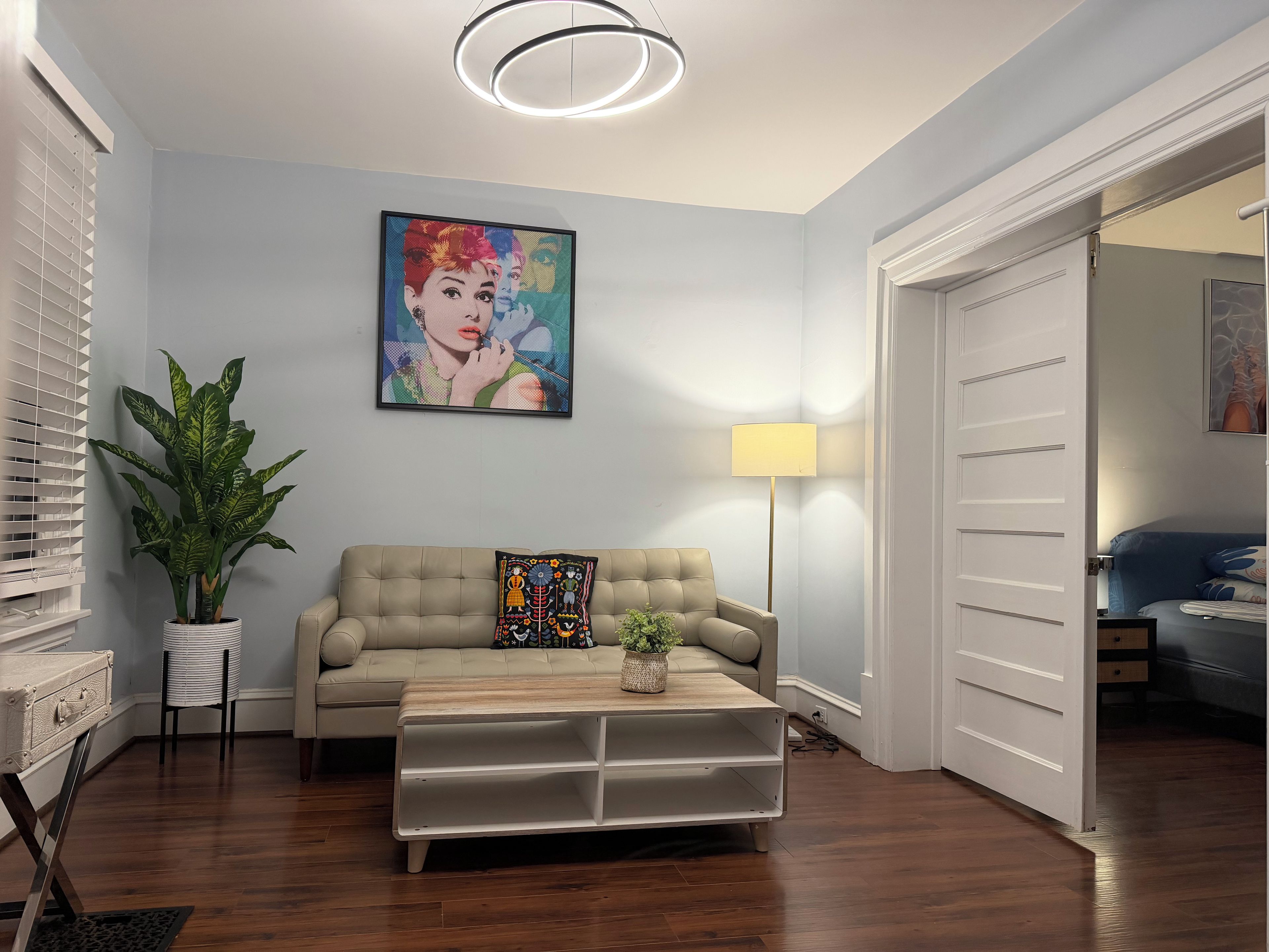 Een woonkamer met een beige bank, een salontafel, een potplant, een schilderij aan de muur en een vloerlamp.