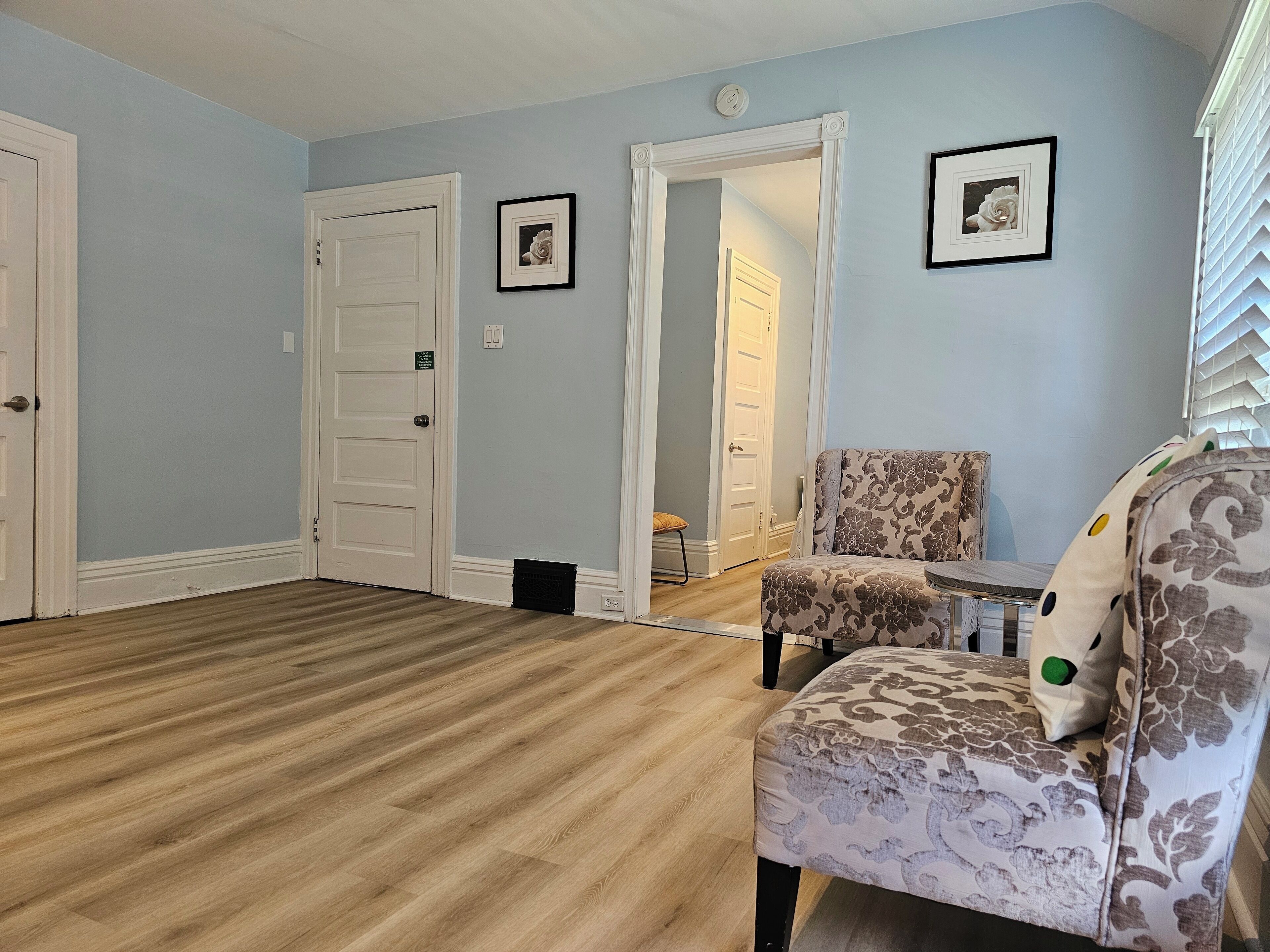 A room with light blue walls, a wooden floor, a patterned armchair, a small table, and two framed pictures on the wall.