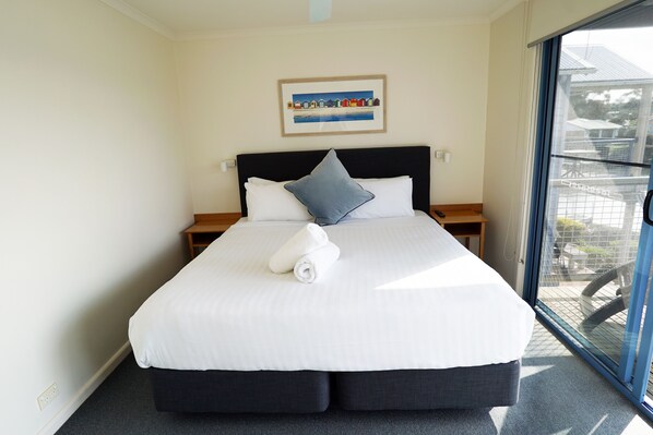 A neatly made bed with white linens and a blue pillow, a framed picture on the wall, and a view through a glass door.