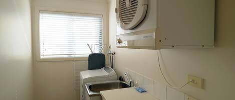 A laundry room with a washing machine, a drying rack, a sink, and a window with blinds.