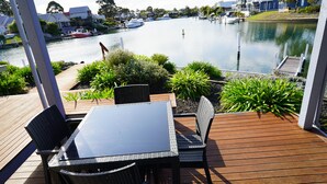 Una terraza con mesa y sillas que da a una masa de agua tranquila, con barcos y casas al fondo.