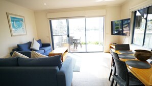 Un salón con un sofá azul, una mesa de comedor de madera con sillas negras, un televisor montado en la pared y un ventanal con vistas al exterior.