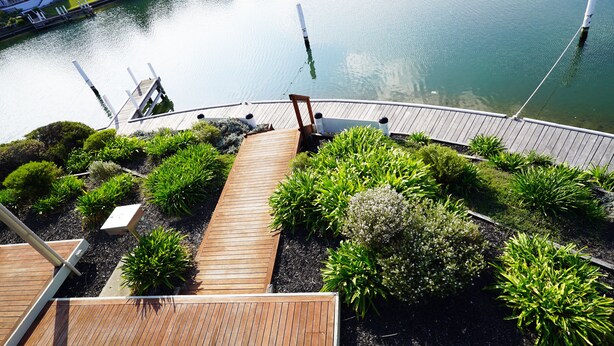 Una terraza de madera con vista a un lago tranquilo, rodeada de exuberante vegetación y un muelle con barcos.